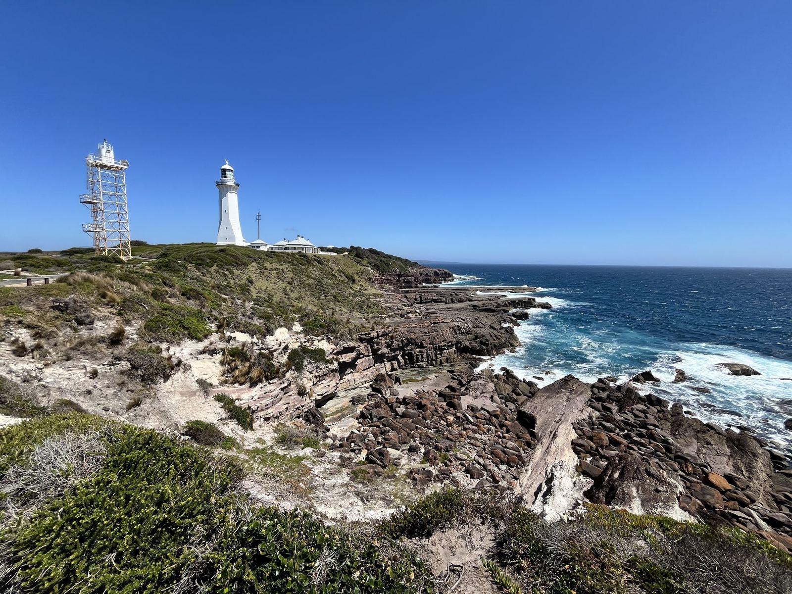 Red-brown layered rock platforms at the end of the headland with the lighthouse and a tower behind, sea breaking below
