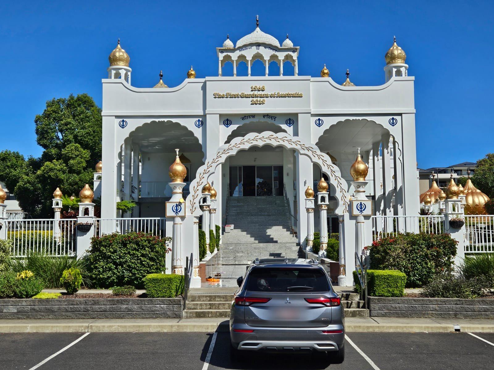 A white gurudwara with gold domes and an arched entrance, its sign reading The First Gurudwara of Australia, a car parked in front