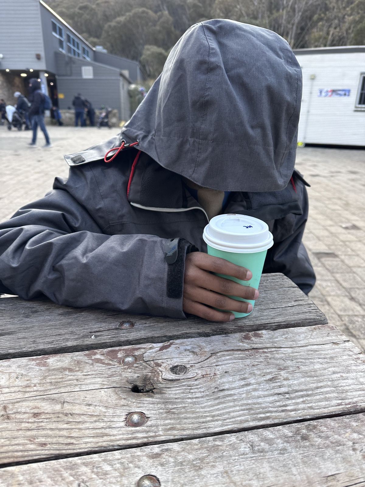 A child in a grey hooded jacket slumped forward over a picnic table with the hood pulled down over their face, both hands around a takeaway cup