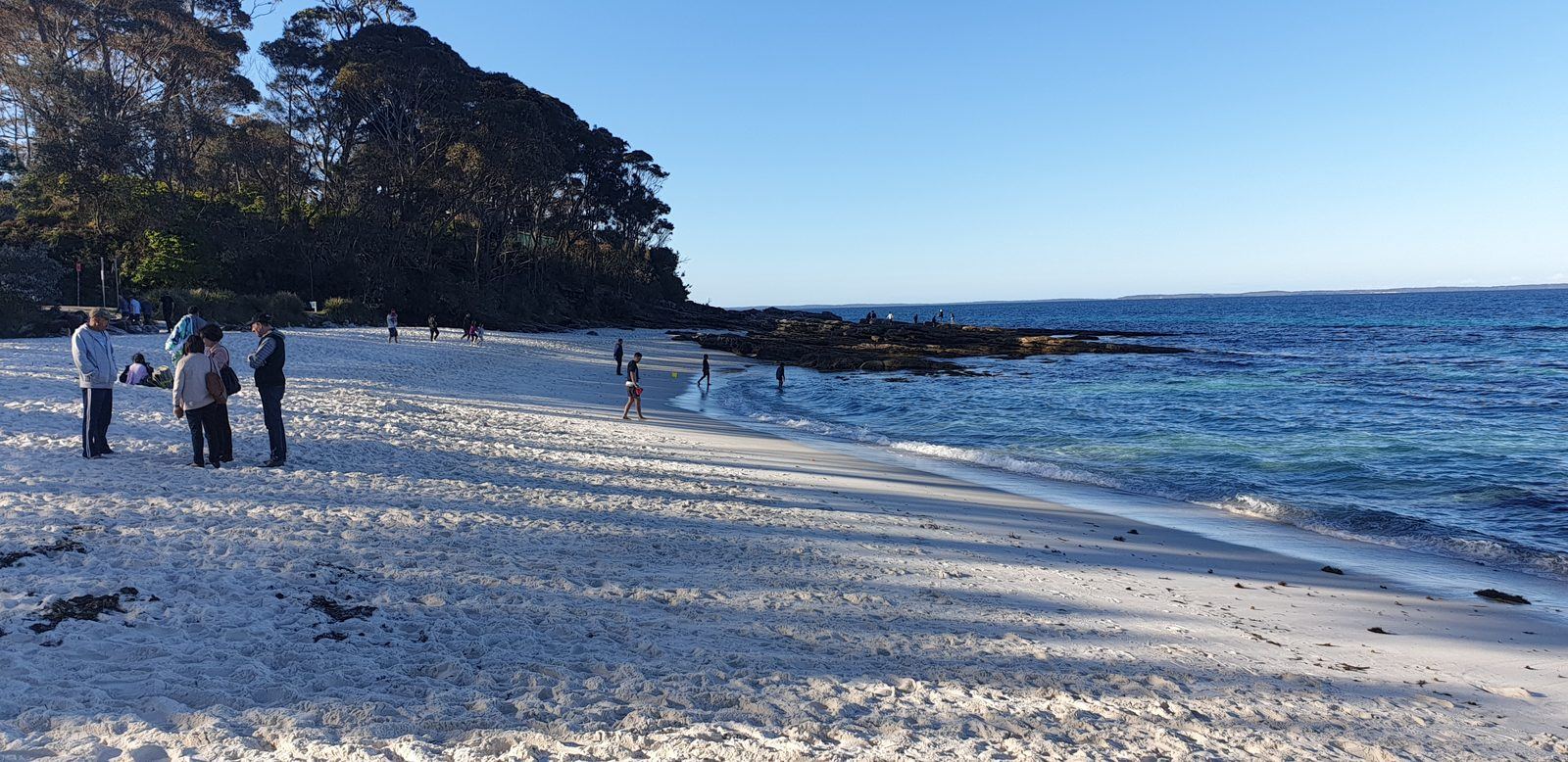 Looking along the white beach from sand level, people standing near the water and a wooded headland running out to the right