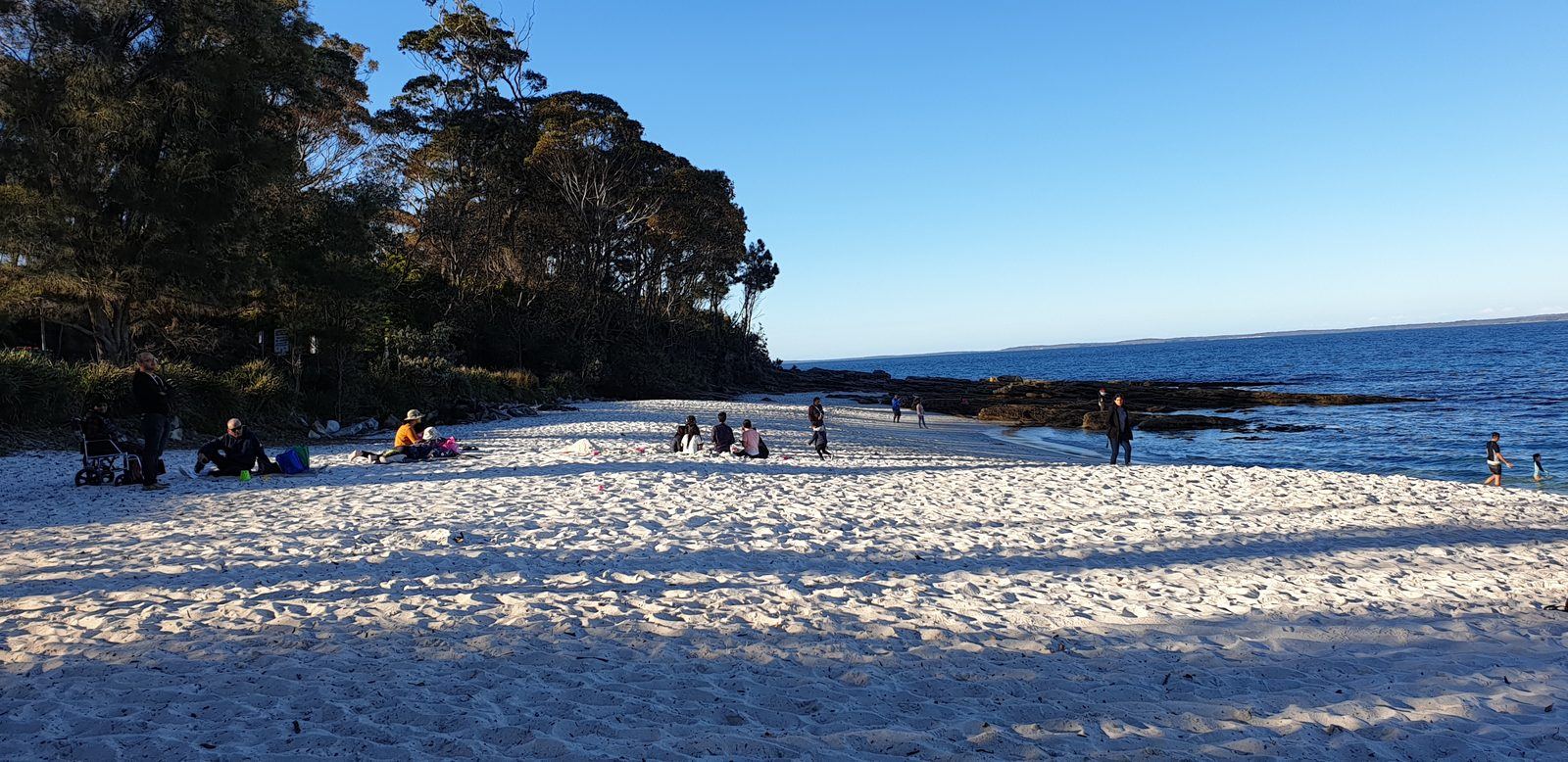 The same beach with long shadows from the treeline falling across the white sand, families sitting near the trees