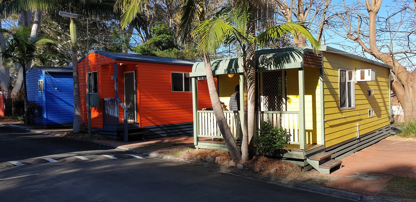 An orange cabin and a yellow cabin with a small covered porch, palms growing between them and a blue cabin further along the road