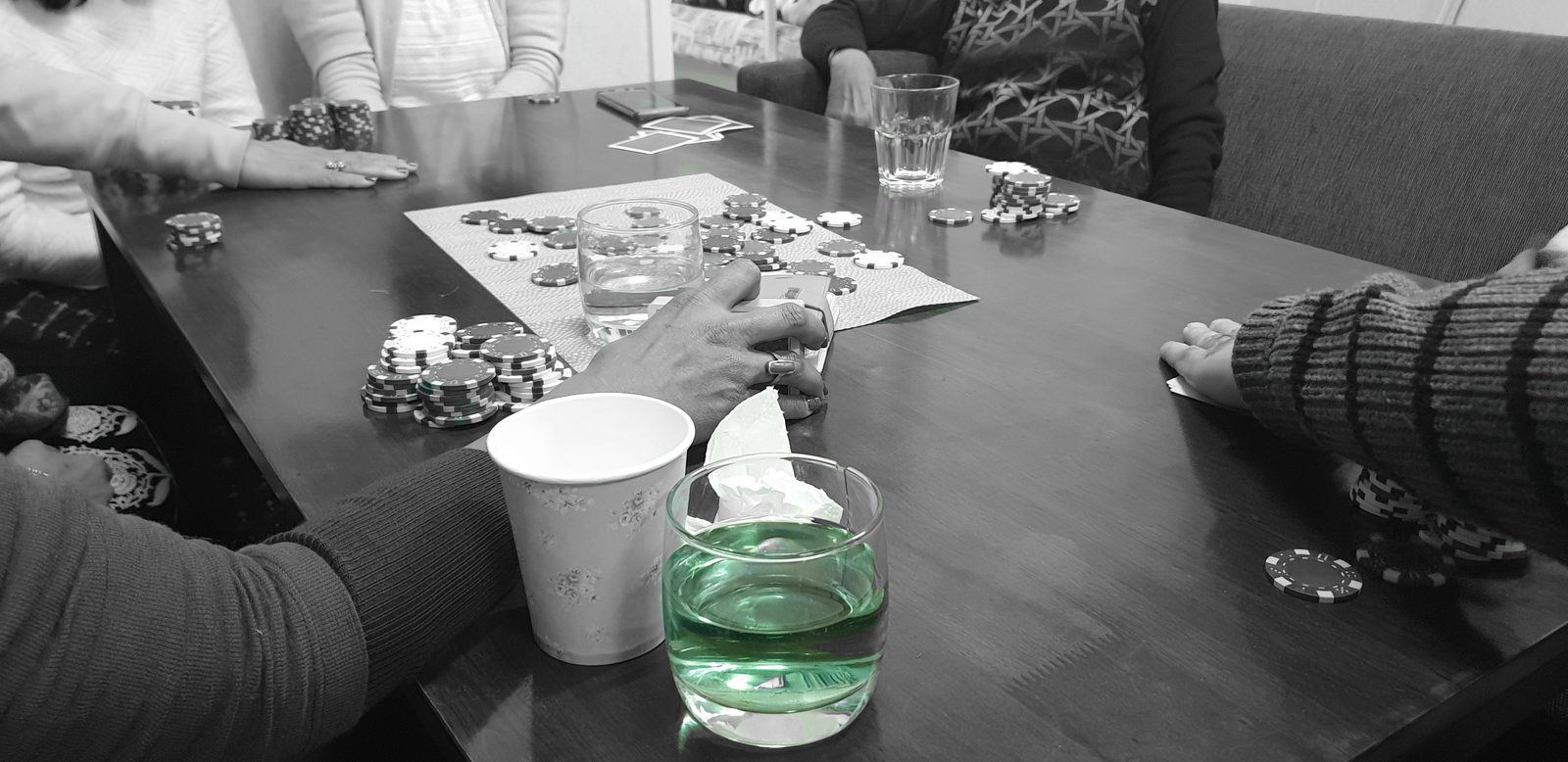 A card game in progress on a dining table inside a cabin, chips stacked around the table and hands reaching in, shot in black and white