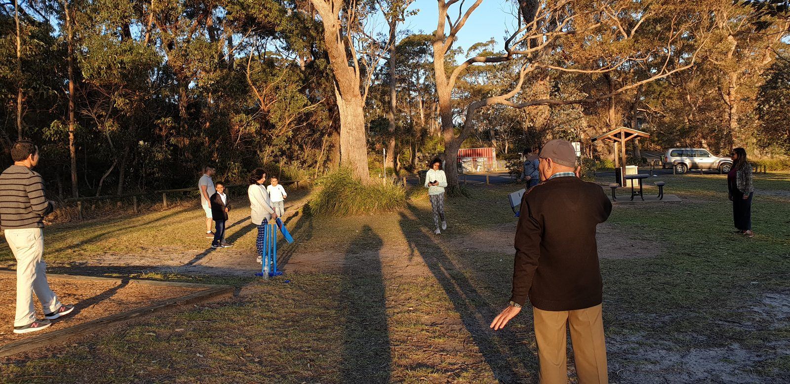 A group playing backyard cricket on the mown grass of the foreshore reserve in low evening sun, long shadows across the ground and gum trees behind