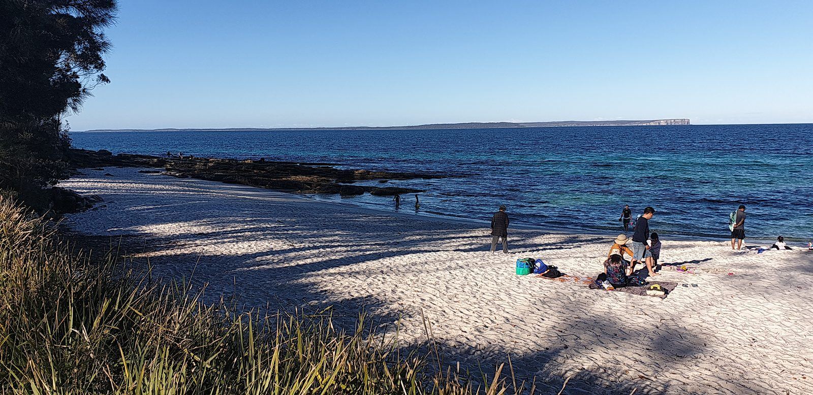 A curve of very white sand with clear pale-green water and a dark rock shelf running out from the headland, bush coming down to the beach