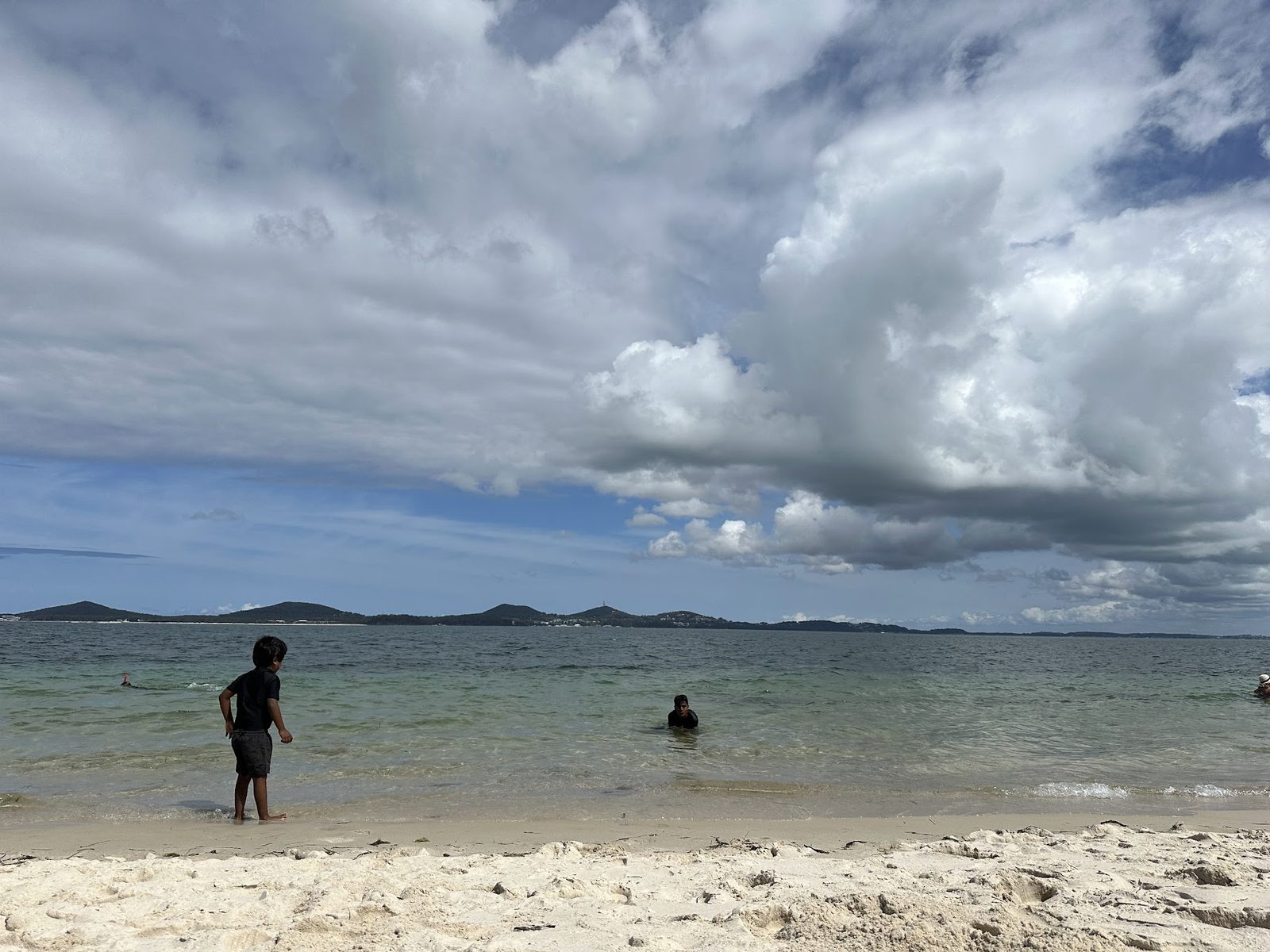 A very shallow clear bay with swimmers standing in knee-deep water, and a run of rounded hills across the water with a mast on the highest of them