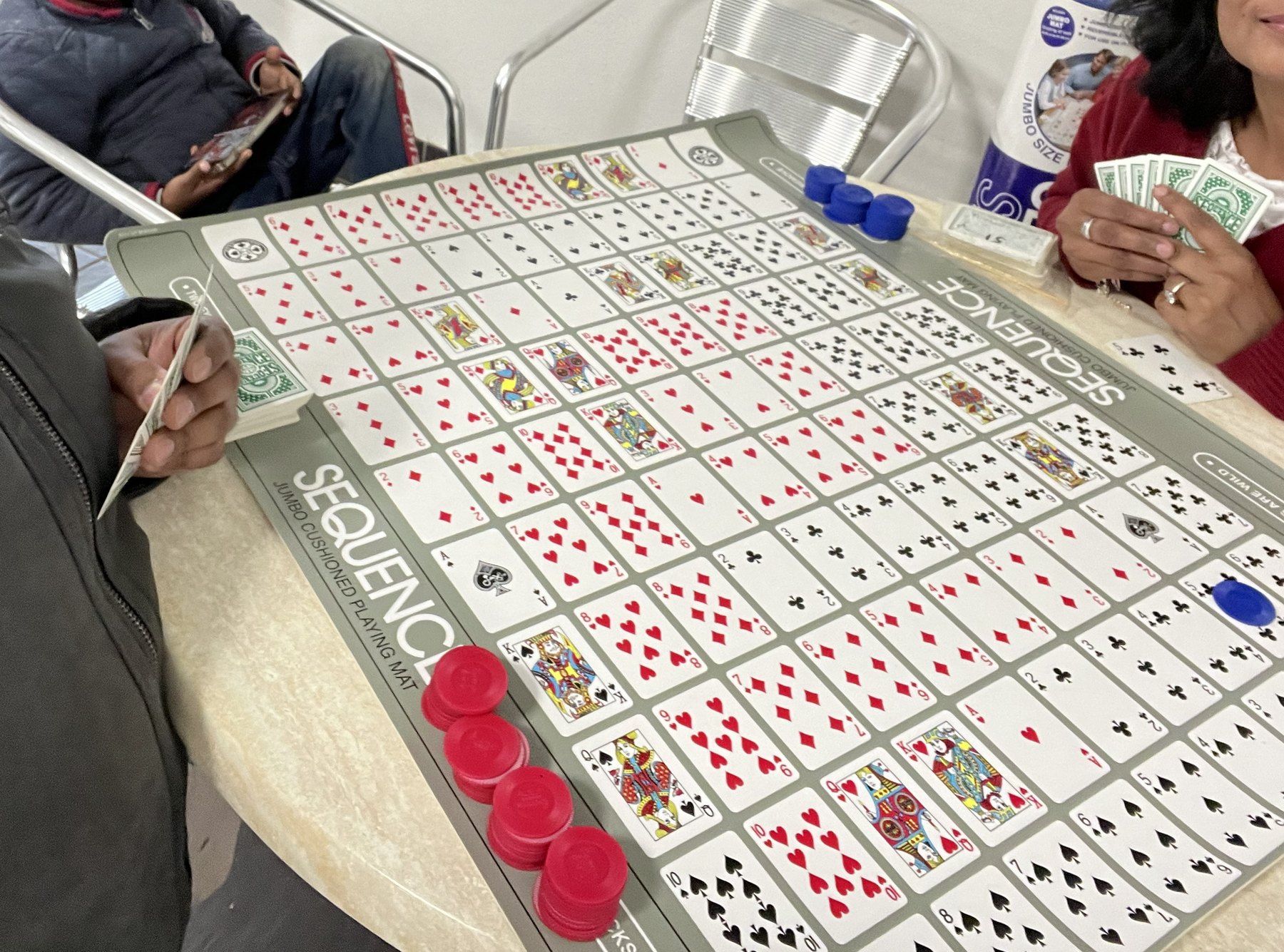 A game of Sequence laid out on a camp kitchen table, hands holding cards around the board and red and blue counters in play