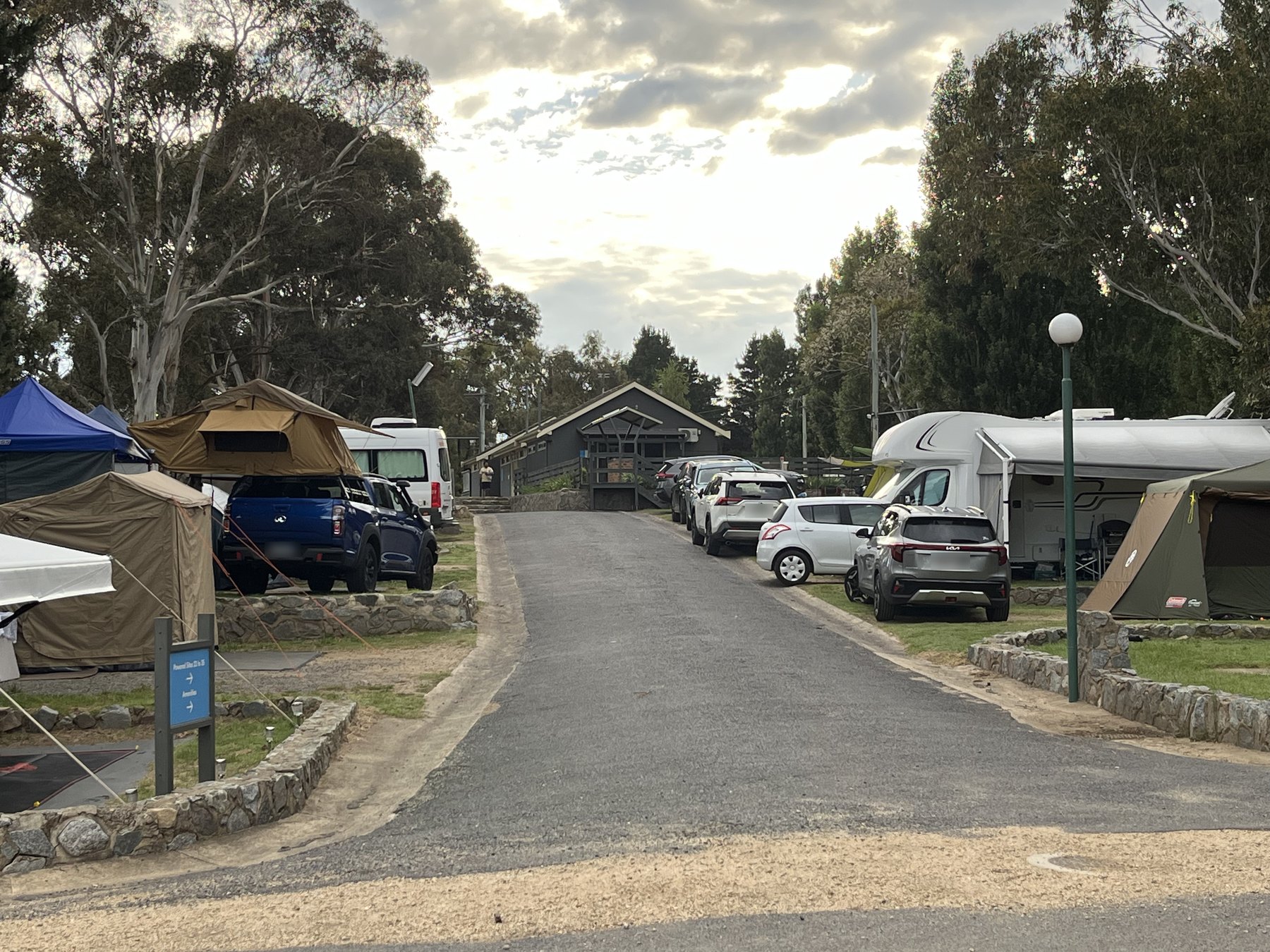 A park road running uphill between pitched tents, a rooftop tent, caravans and parked cars towards a timber amenities building in early light