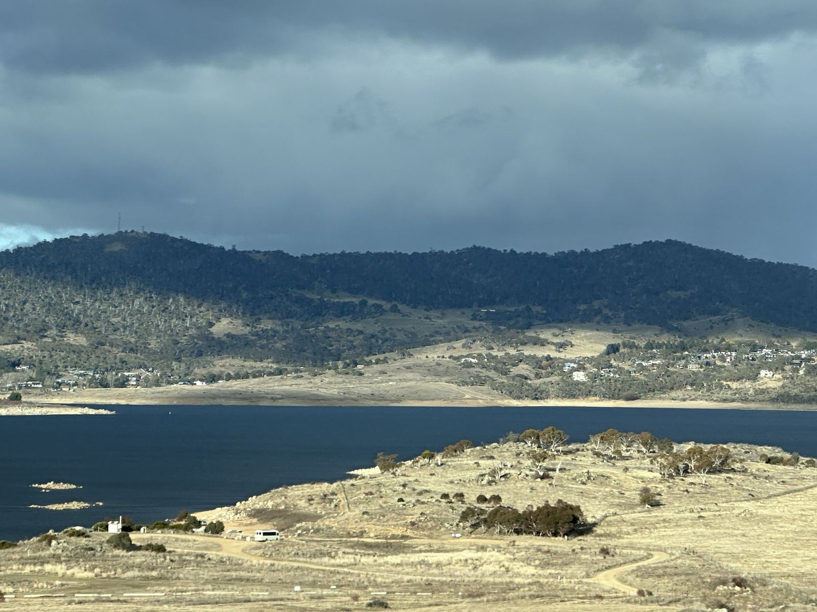 Lake Jindabyne under heavy cloud, with the town on the far shore and dry winter grass in the foreground