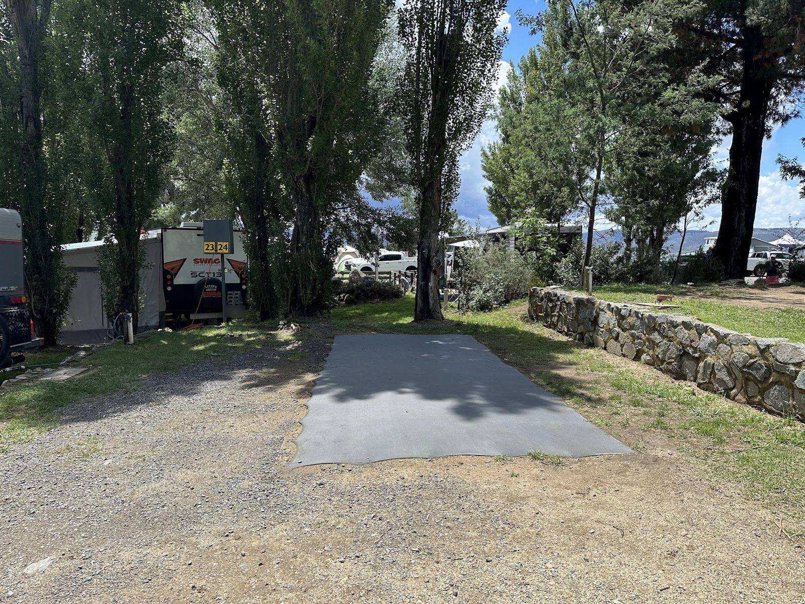 An empty numbered concrete camp slab with a low stone retaining wall and poplars behind, everything packed away, a neighbour’s camper trailer on the next site