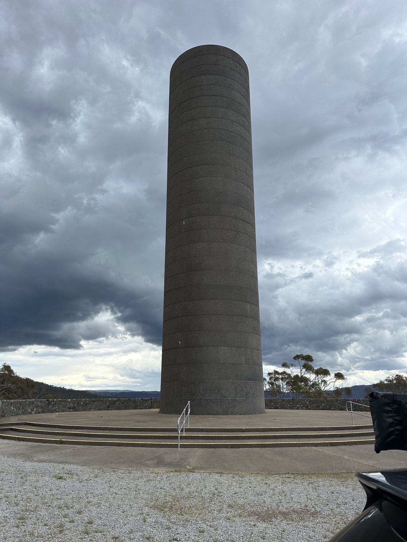 A tall ringed concrete cylinder on a circular platform with steps, a stone wall and a ridge behind, under dark storm cloud