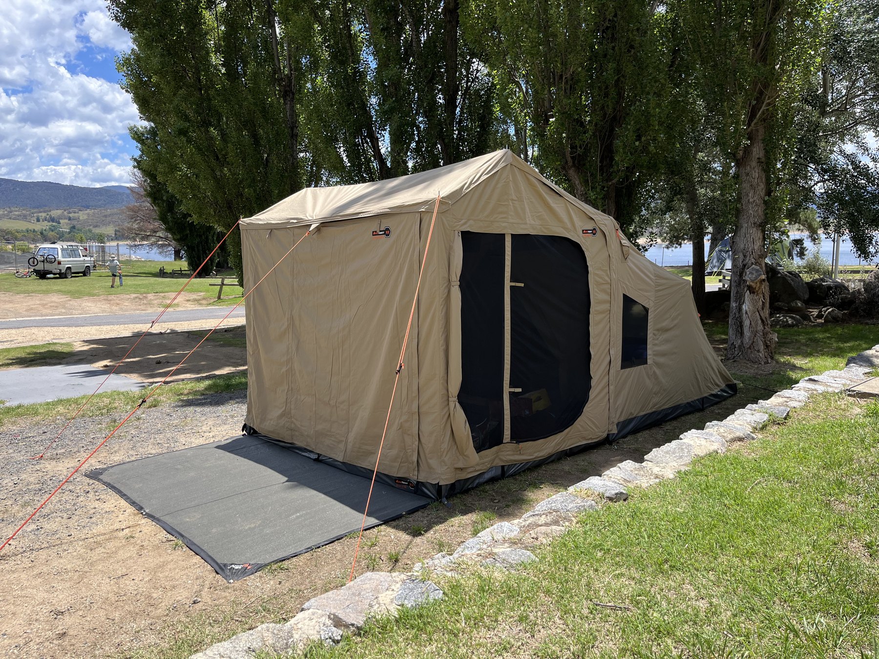 A canvas tent with its front room zipped shut on a gravel and grass site, a ground mat in front, poplars and the lake behind
