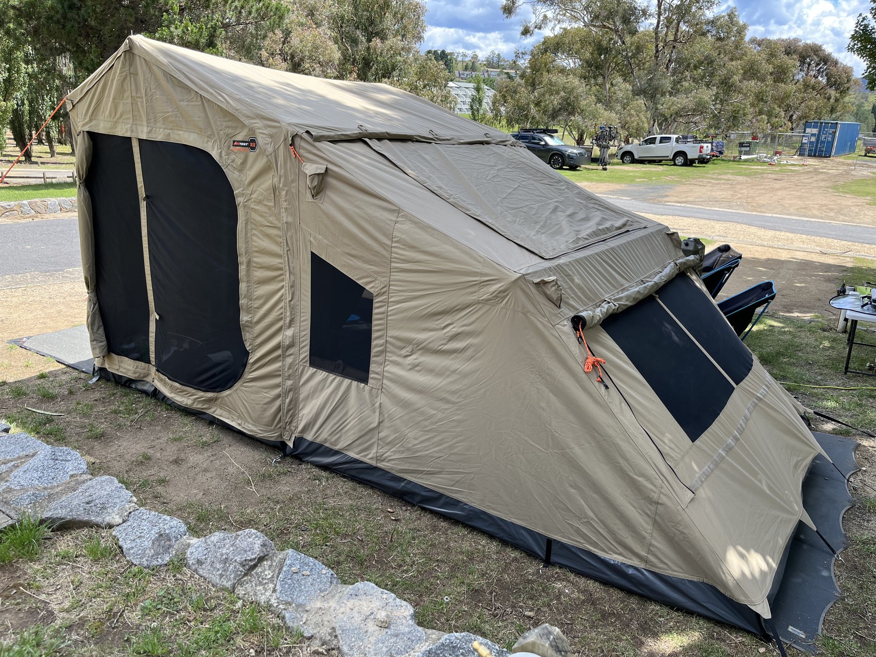 The side of the same canvas tent, its guy ropes pegged into dry ground, camp chairs and a table beyond it and cars parked across the park road