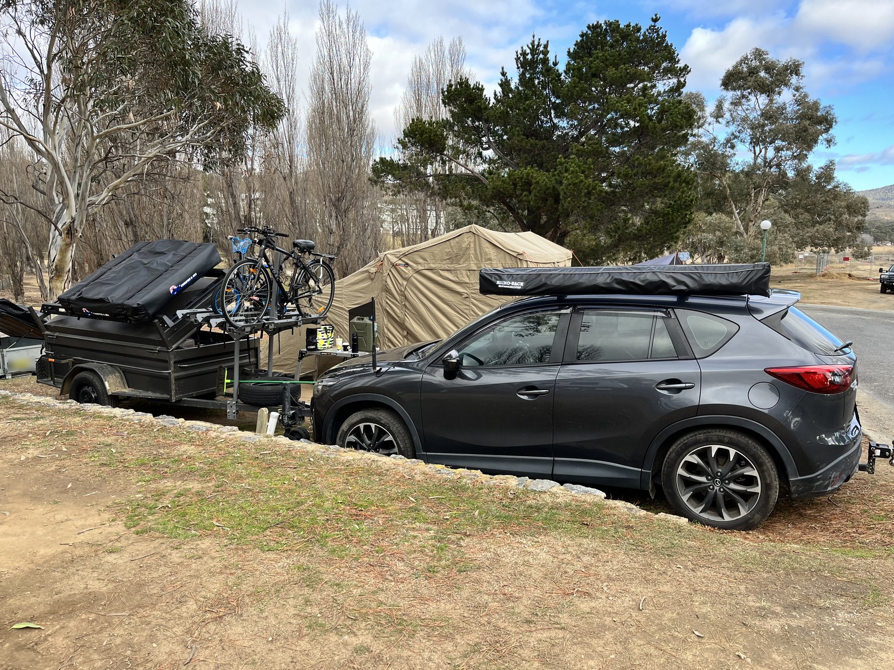 A grey SUV with a roof awning hitched to a box trailer carrying bikes, parked beside a canvas tent on the same grassy site under bare poplars