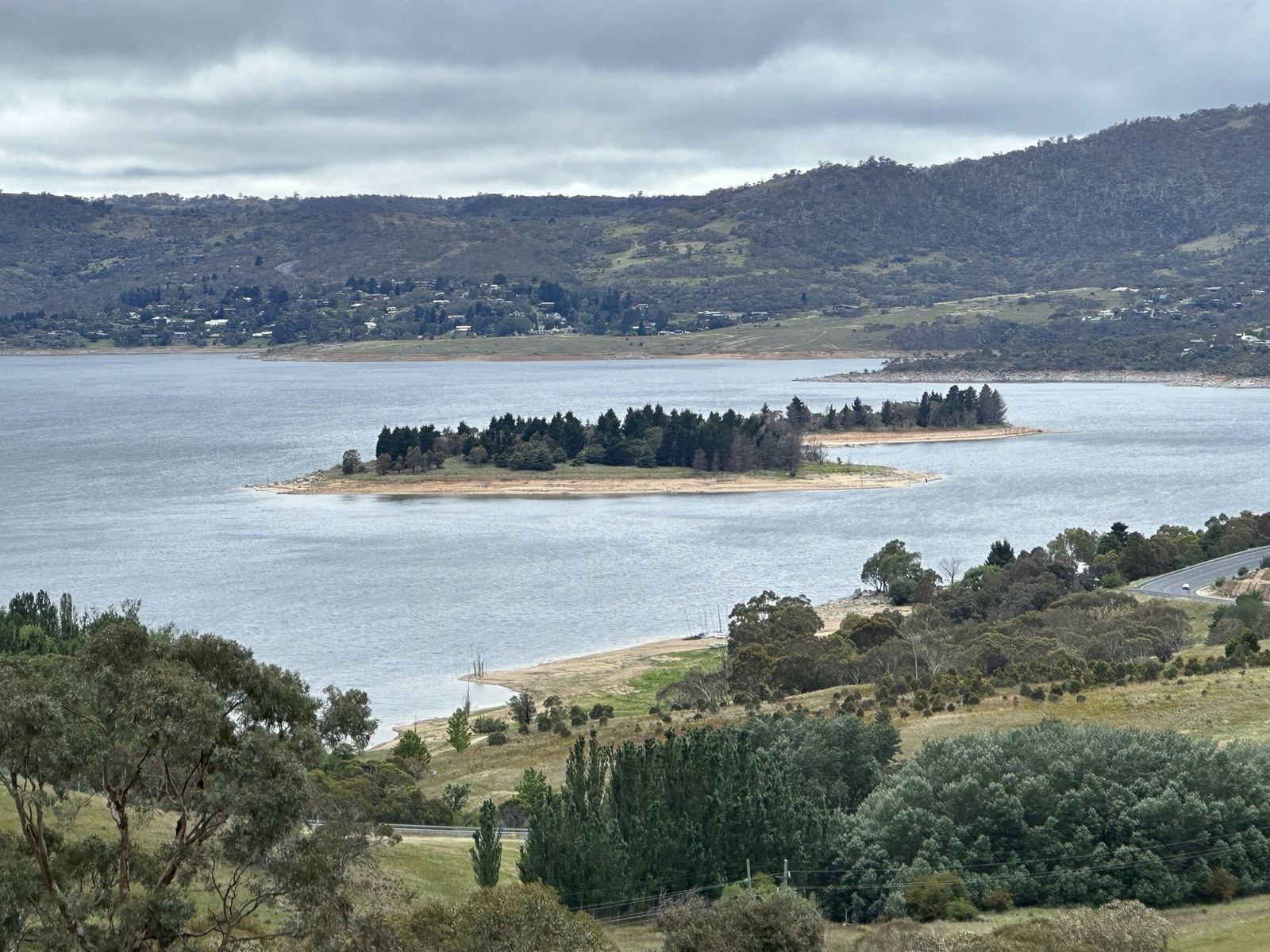 A wide lake seen from a hillside, with a wooded island in the middle distance and houses along the far shore