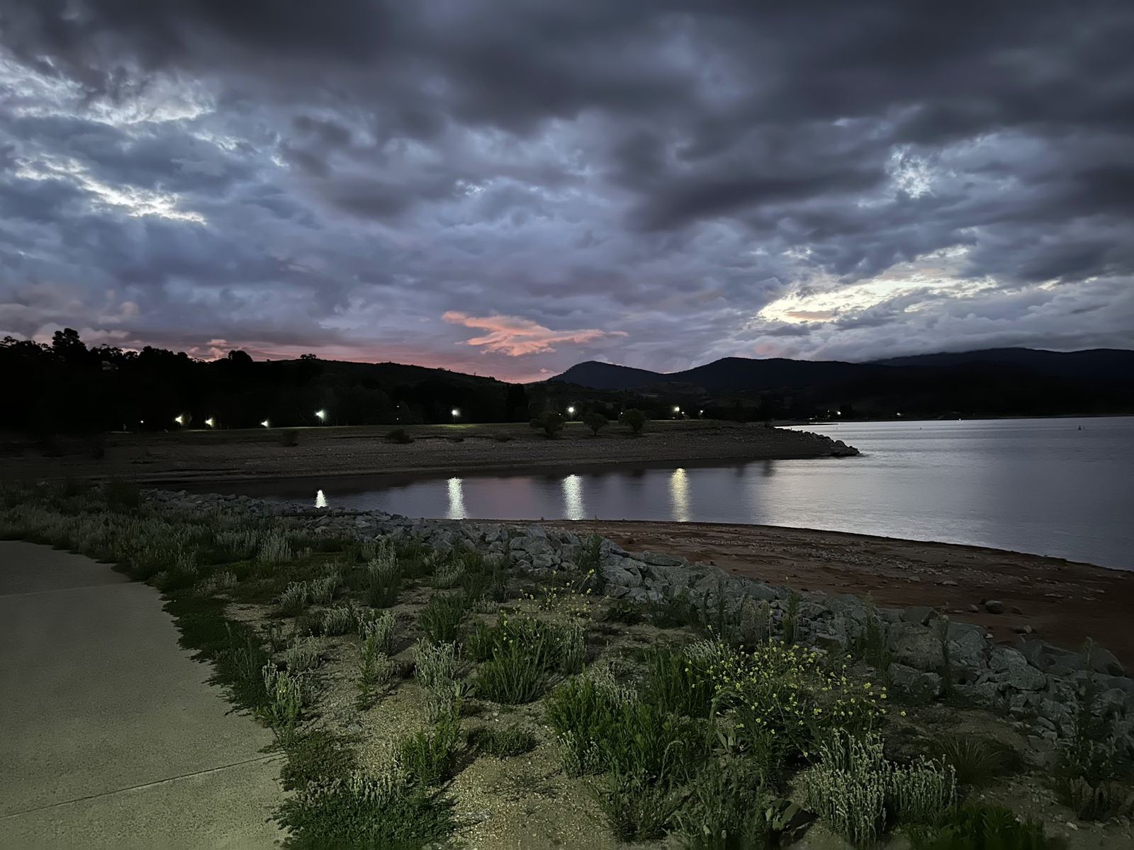 Lake Jindabyne at dusk, pink cloud over dark hills and lamps along the shore reflecting in still water