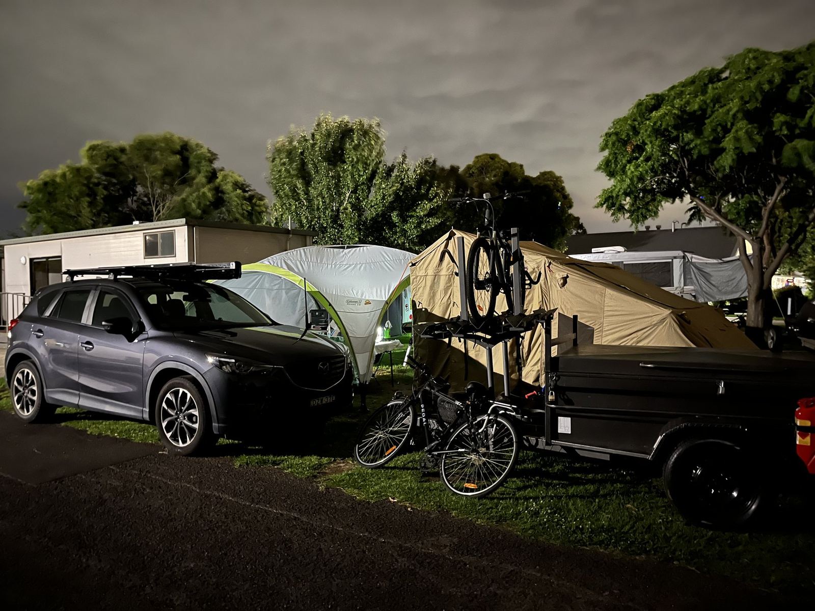 The car, trailer and bikes beside the tent in the dark, cabins and vans of the park behind