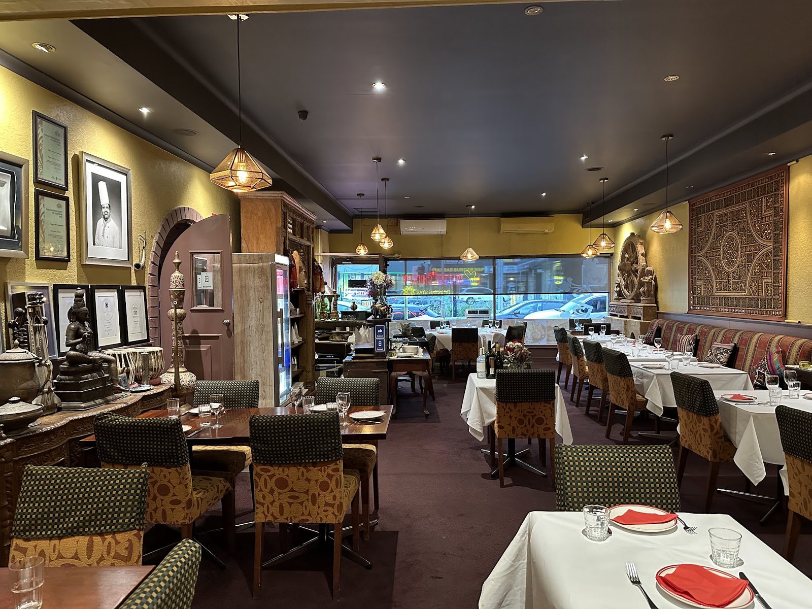 The dining room of an Indian restaurant, empty tables laid with white cloths, a carved stone wheel and framed certificates on the wall
