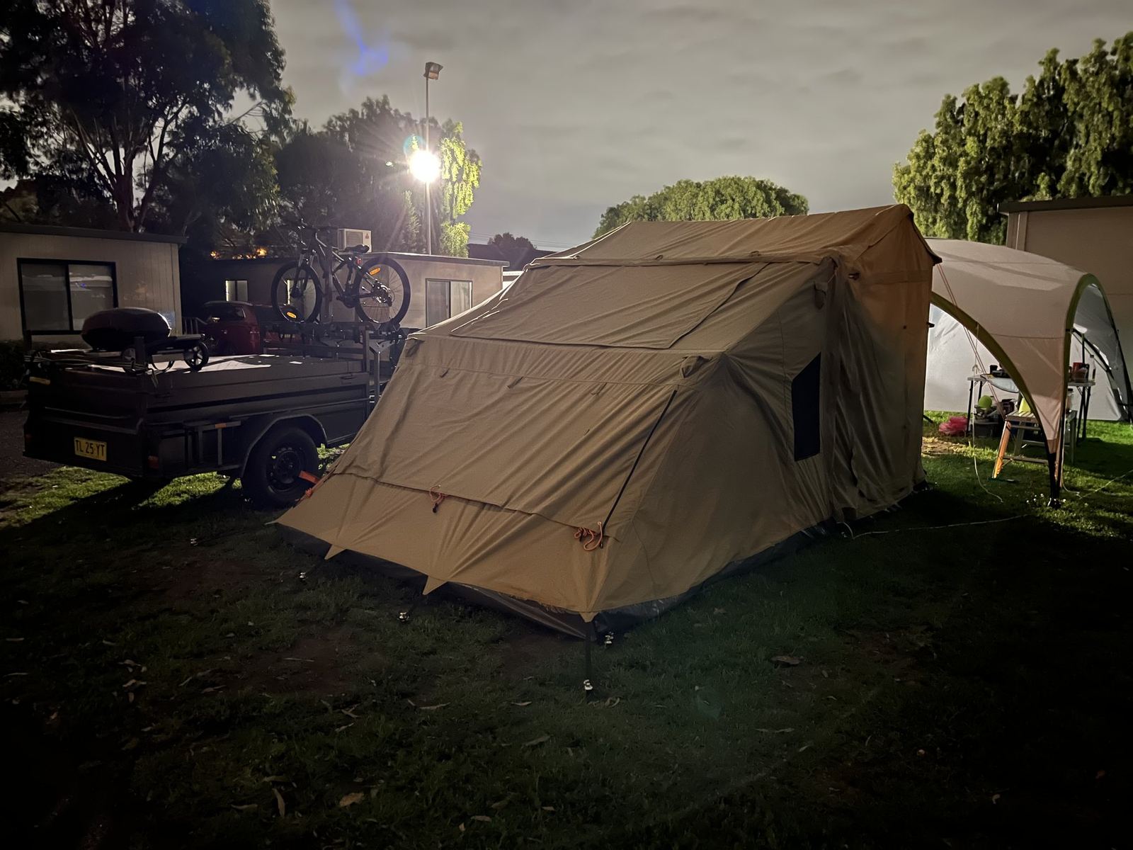 The canvas tent pitched on grass at night, lit from one side, with a shade shelter behind and a floodlight through the trees