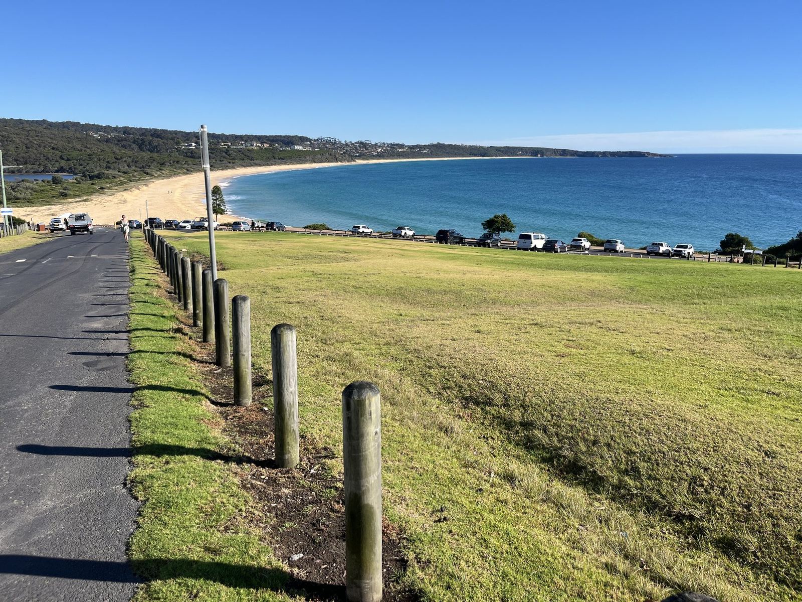 A long curve of beach seen from the grassy verge above the road, blue water, a wooded headland at the far end and cars parked along the foreshore