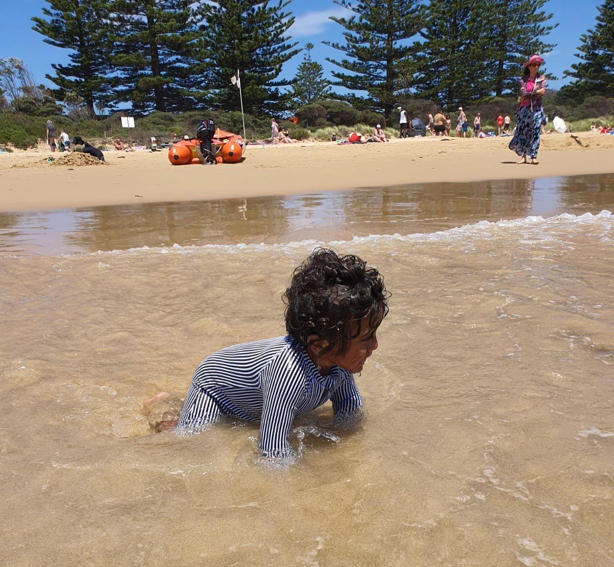 A toddler in a striped rash vest crawling in warm ankle-deep water at the edge of a lake beach, tall Norfolk pines and people on the sand behind