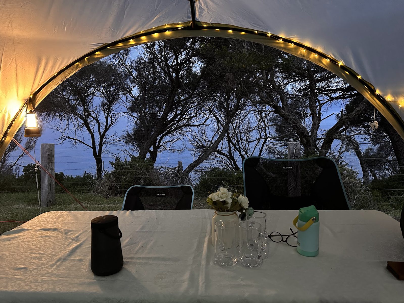Looking out from inside a shade tent at dusk, fairy lights along the arch, a lantern hanging at the side, a laid table and two camp chairs facing the sea through banksias