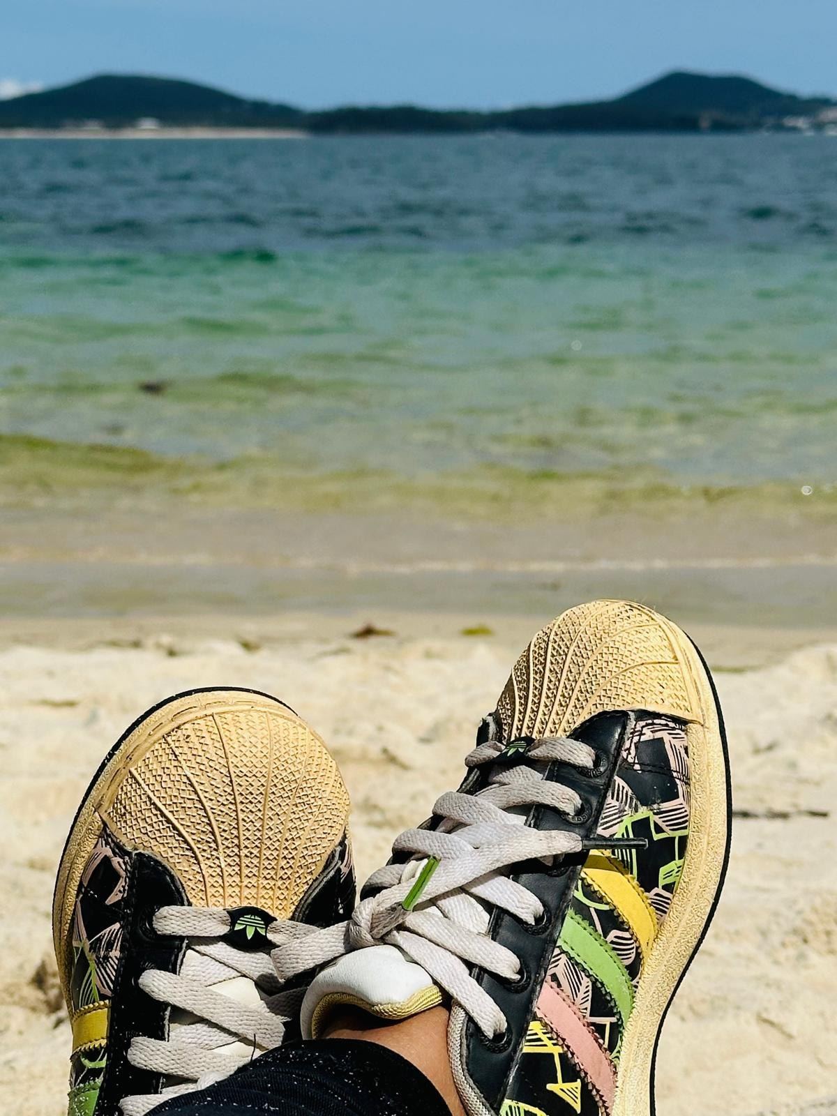 A pair of shoes propped on the sand at the water's edge, clear shallow water and a wooded shore beyond