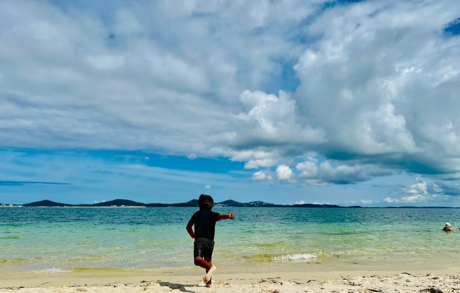 A child running down the sand into the shallow lake water, low hills across the broadwater and big cloud above