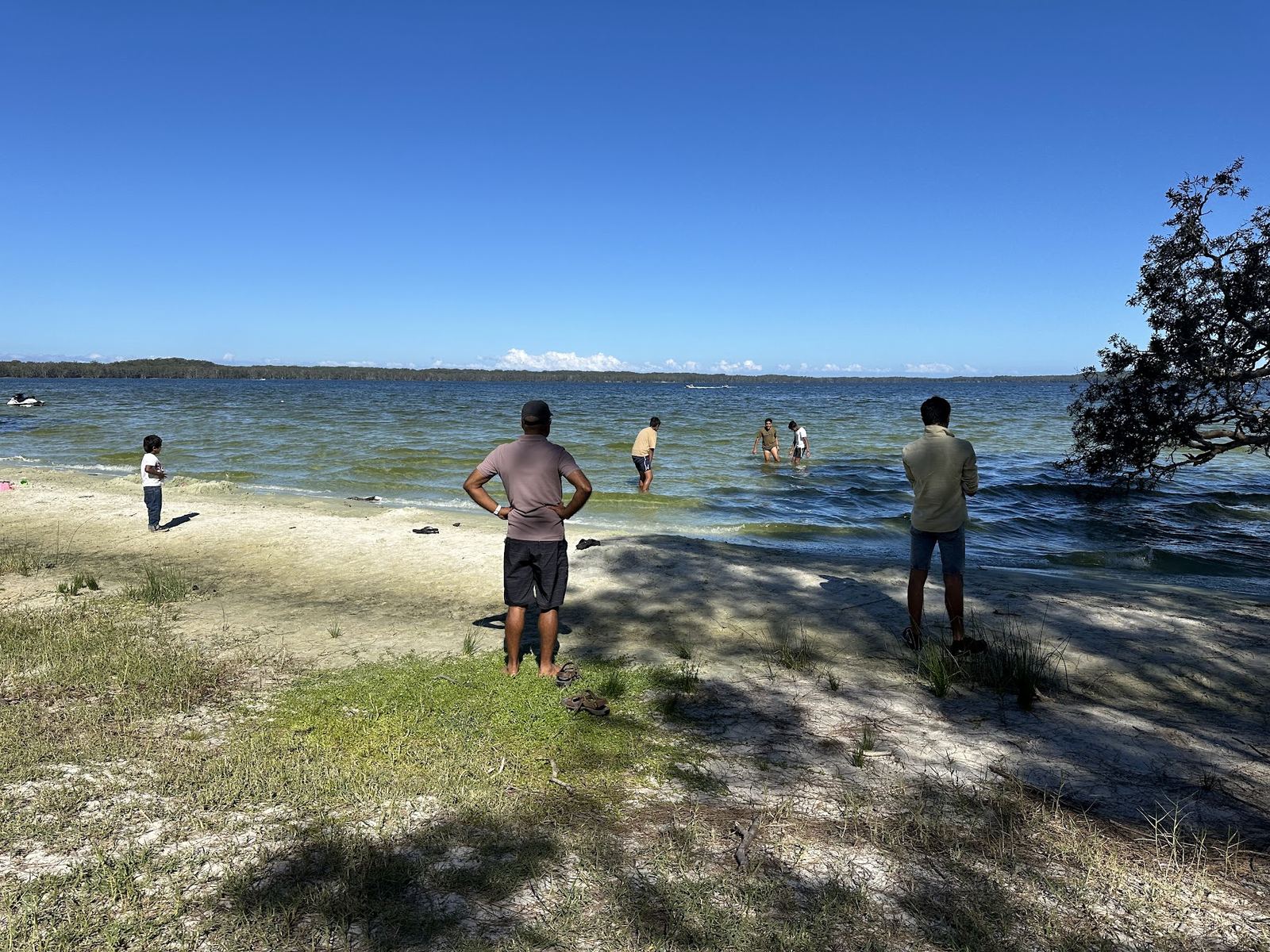 A sandy lake beach with people swimming in the shallows and a jet ski moored offshore, forested shoreline running away on the far side