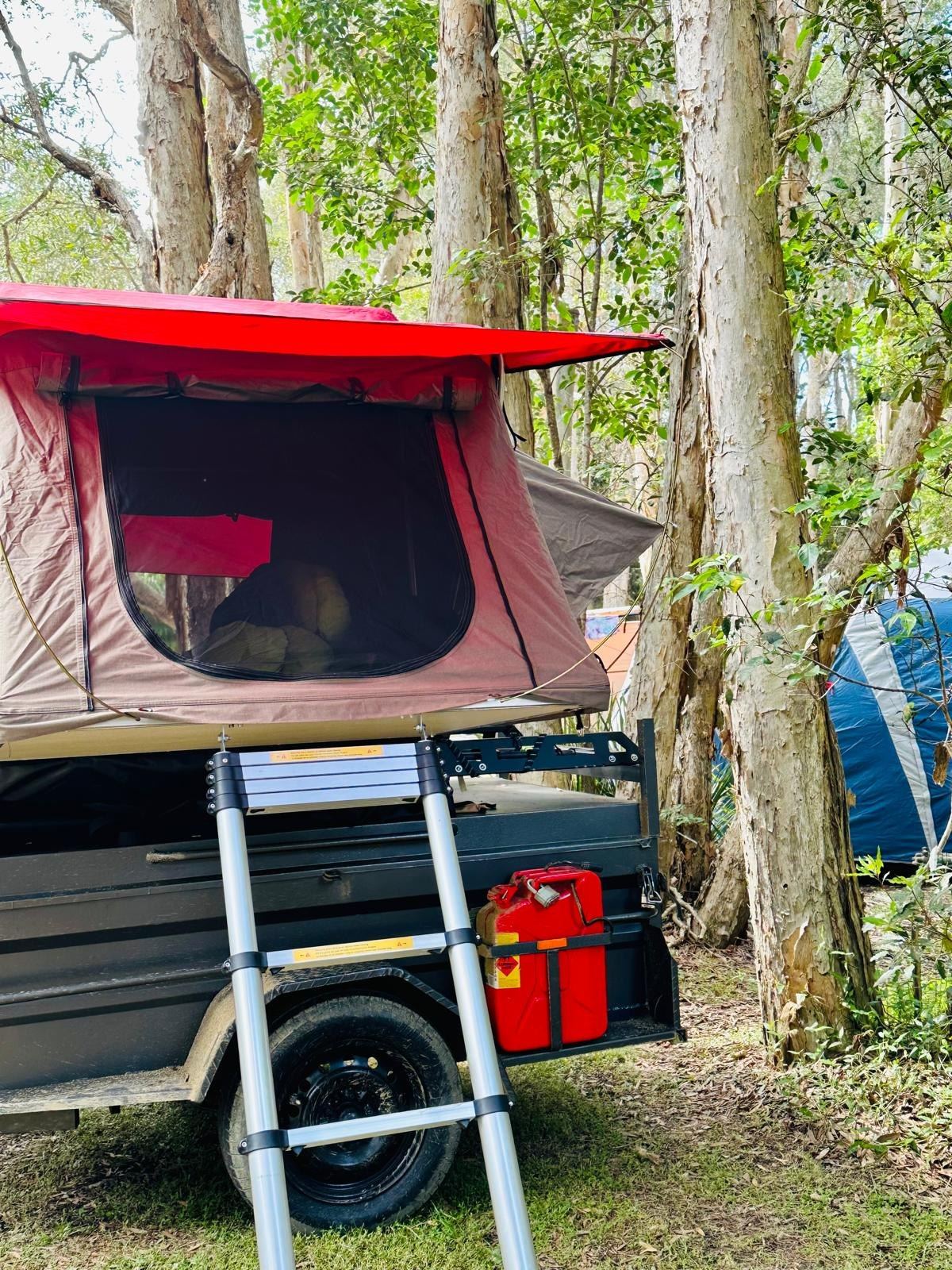 The rooftop tent open on its rack among tall trees, its red rainfly extended over the window