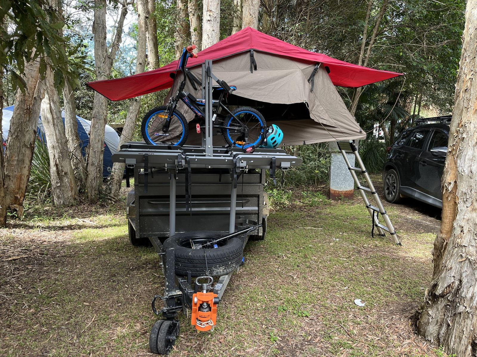 A box trailer with a red-and-tan rooftop tent opened out on a rack above it, ladder down, two bikes carried on a rack at the front