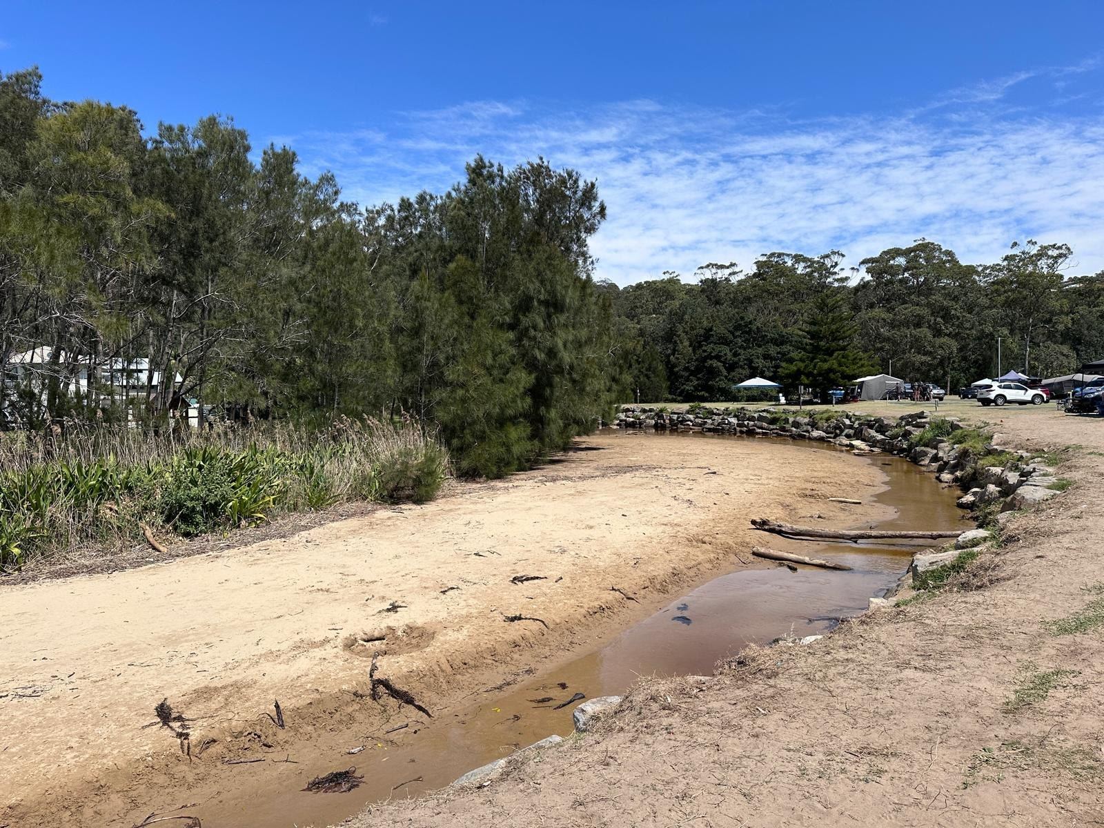 The creek winding through the park between a rock wall and a sandy bank, campervans and tents visible under the trees