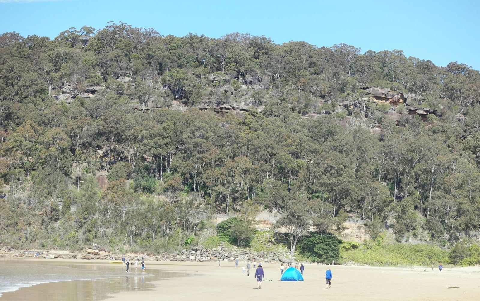 The wooded headland at the end of the beach with a few small figures and a beach tent on the sand below