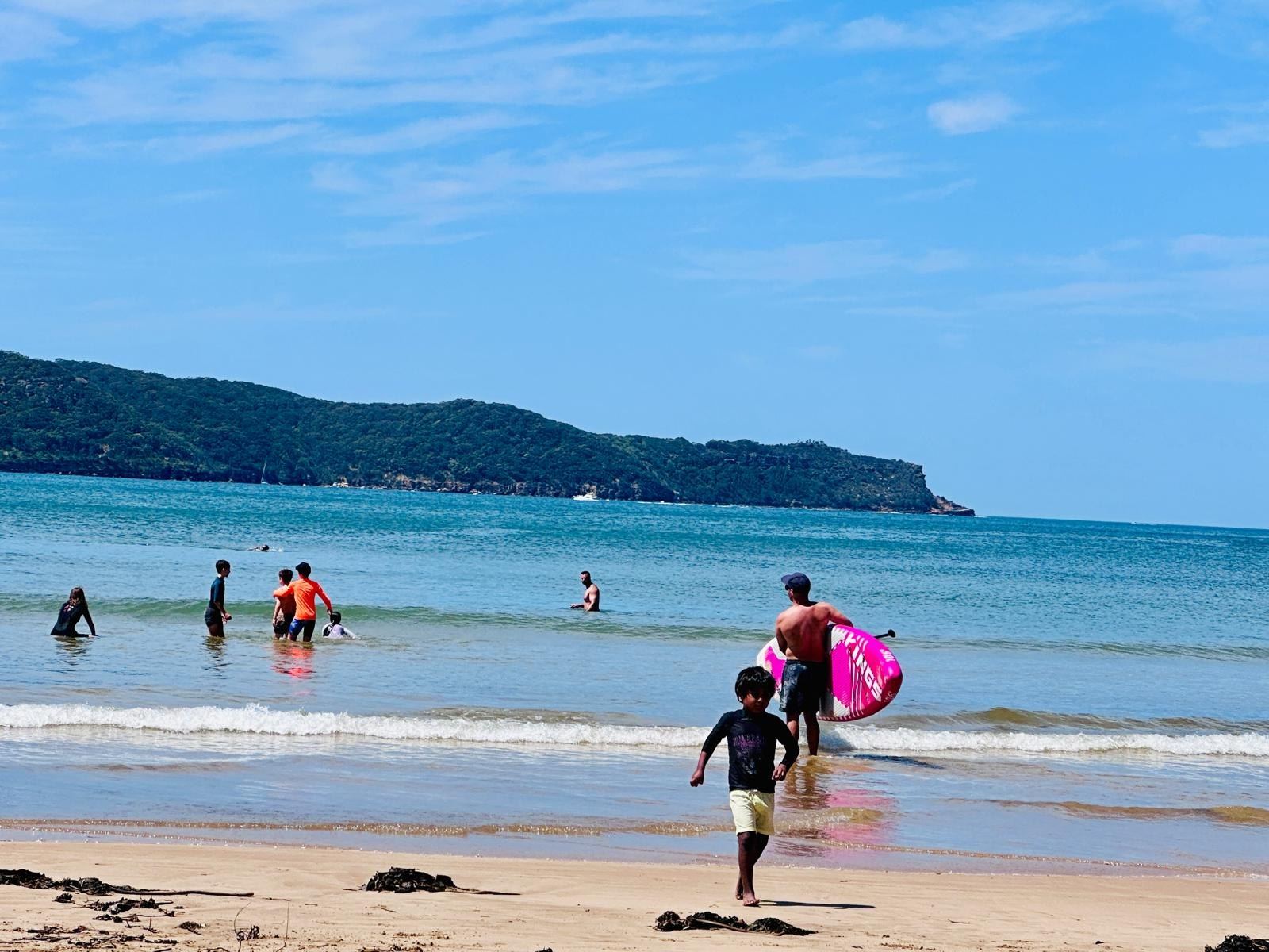People standing and playing in the shallows on a bright day, a child carrying a pink inflatable ring up the sand