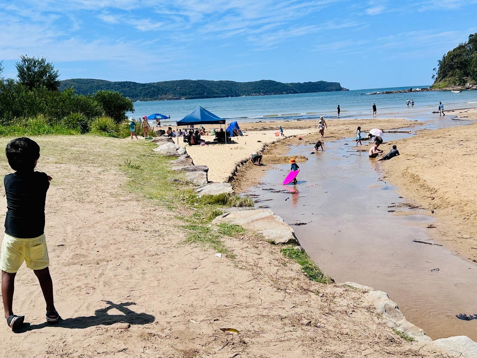 A shallow creek running across the sand to the sea, a child standing on the bank watching, beach umbrellas along the shore