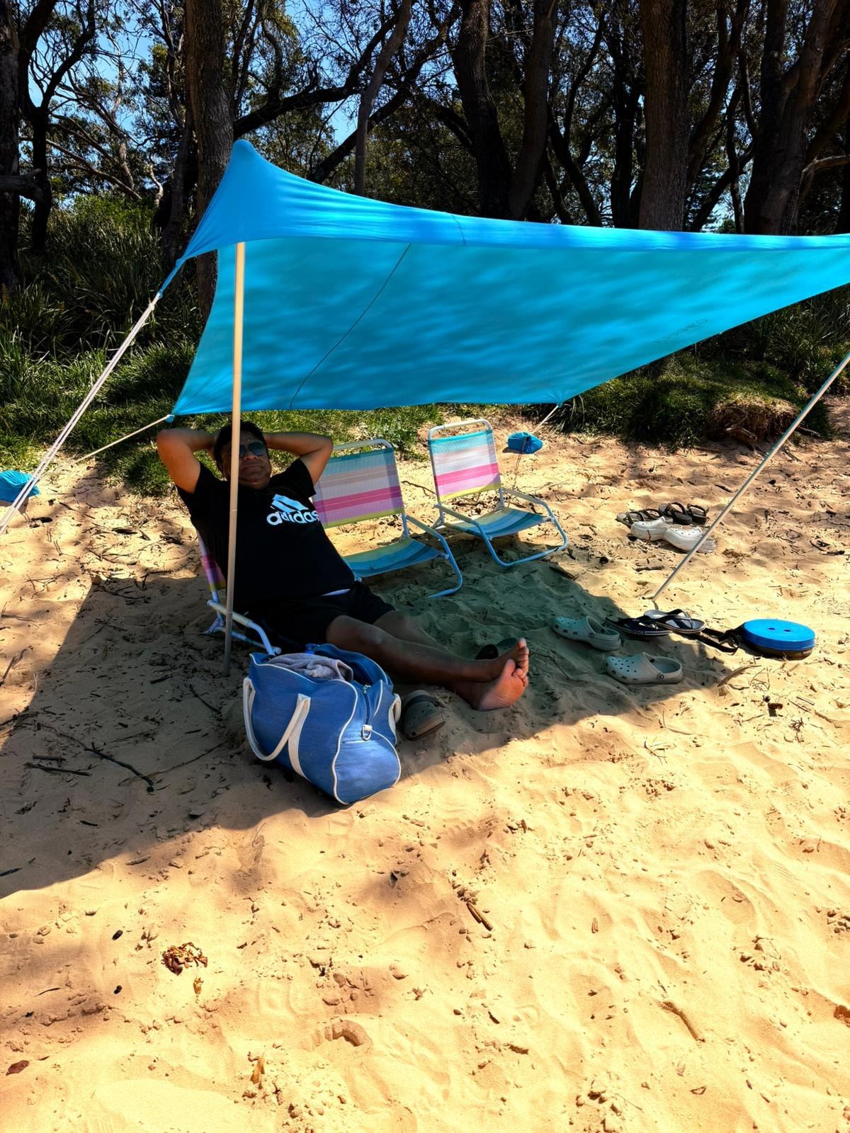 A blue shade sail rigged over the sand with striped beach chairs and bags underneath, trees behind