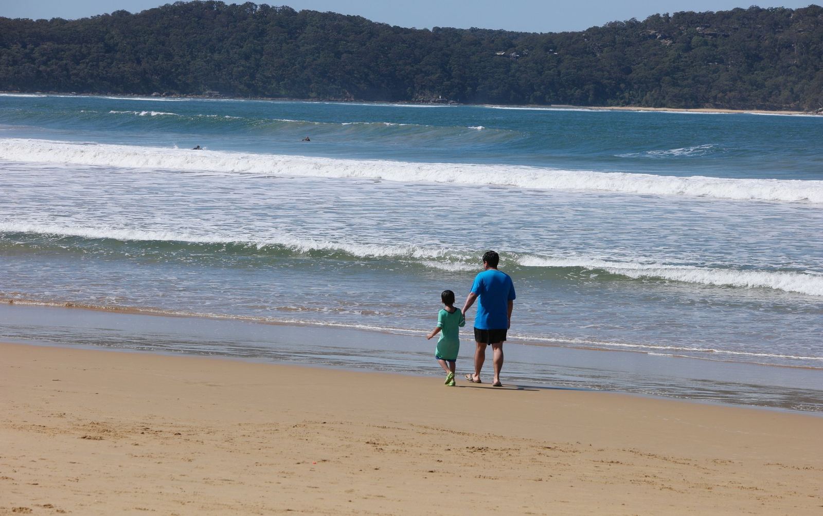 An adult and a small child walking along the wet sand at the shoreline, seen from a distance with surf behind them