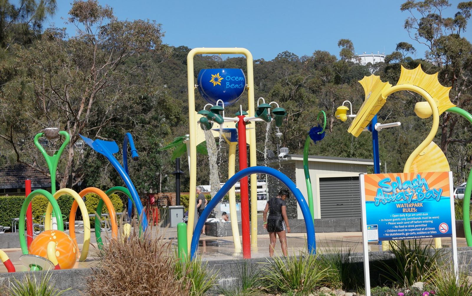 The park's water play area, brightly coloured splash structures and tipping buckets on a paved apron under tall trees