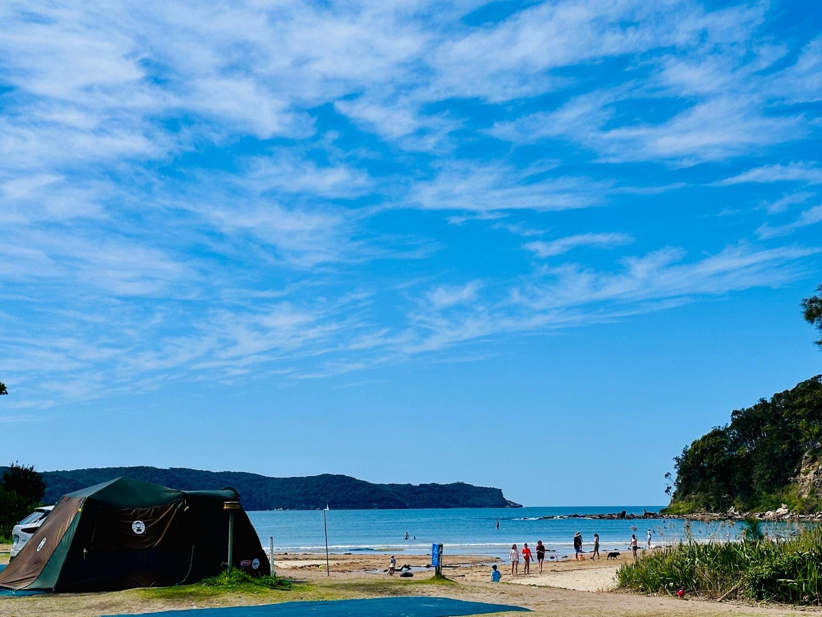 A dark canvas tent in the foreground with the bay, the beach and a wooded headland stretching away behind it
