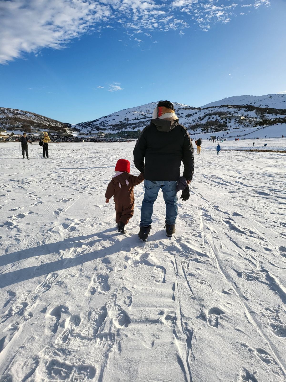 An adult and a small child seen from behind, hand in hand, walking away across tracked snow towards low snow-covered hills