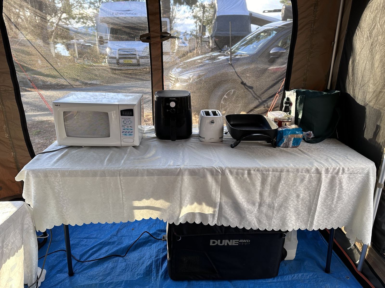 The front room of an Oztent RX-5 set up as a camp kitchen, with a microwave, air fryer, toaster and electric frypan on a covered table and a fridge underneath