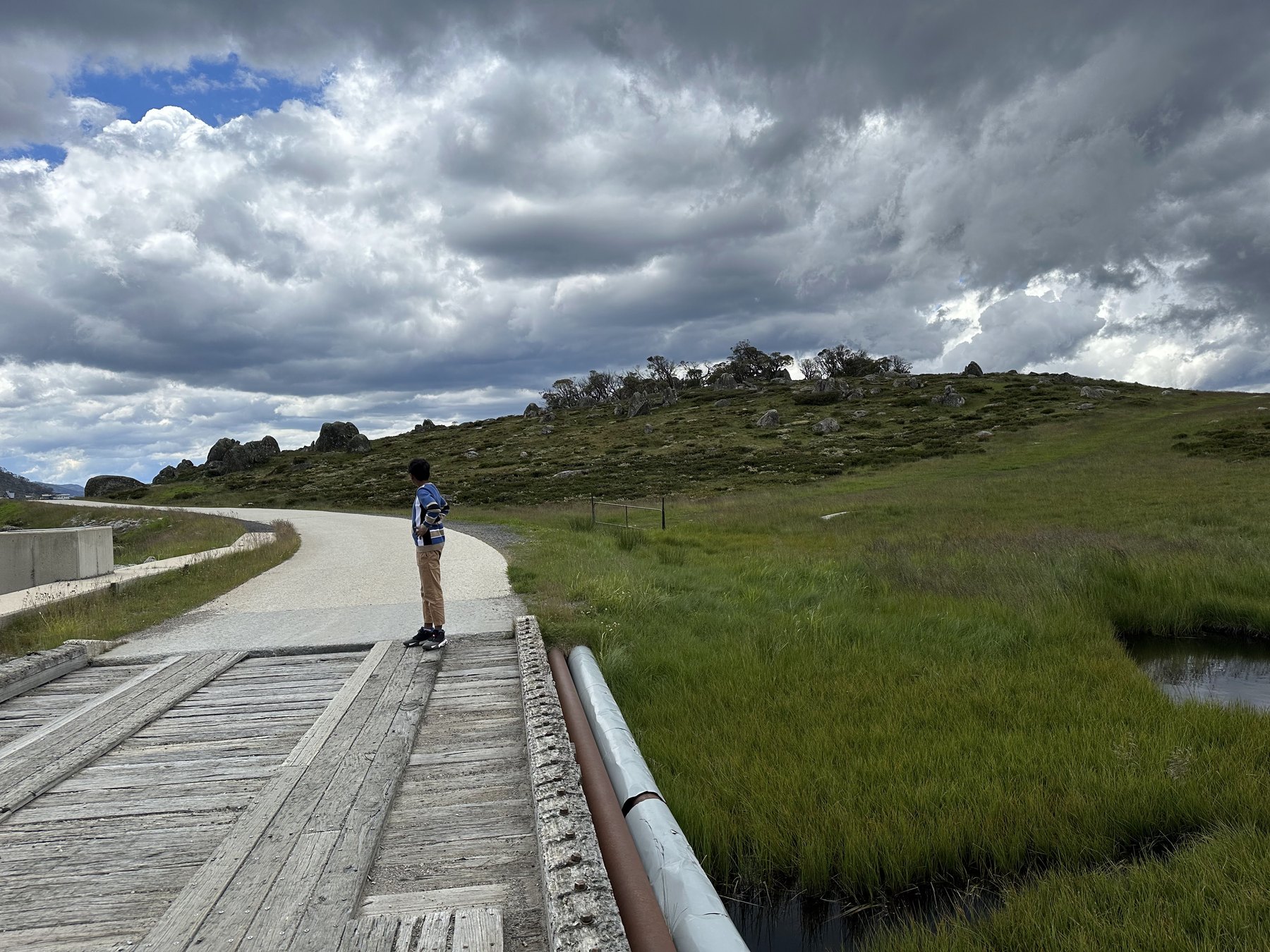 A boy standing on an old timber bridge over a grassy creek, a gravel path curving past a rocky hill under dark cloud