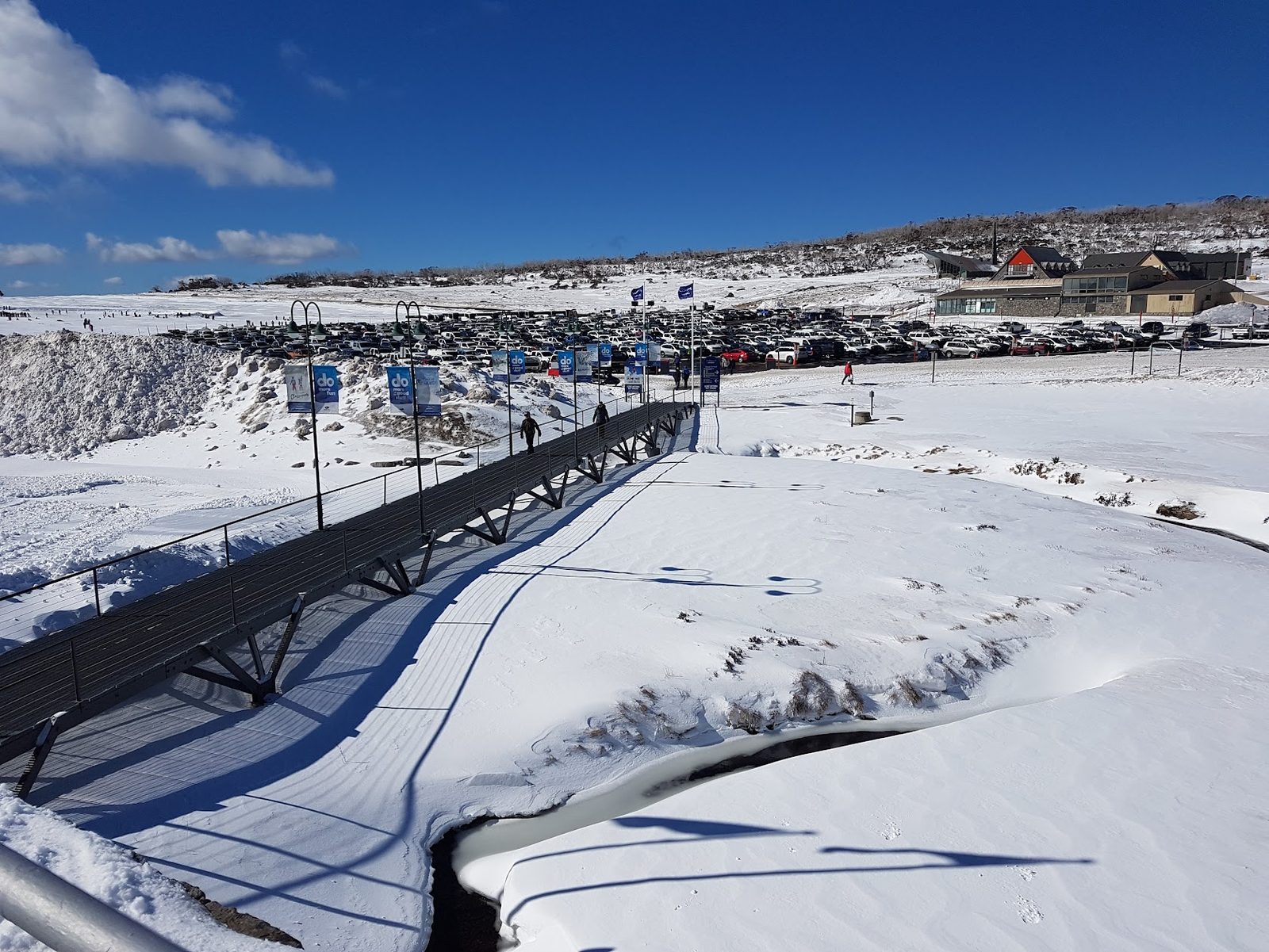 A long steel footbridge over a snow-covered creek leading to a very large car park, banners along the rail and the village buildings beyond