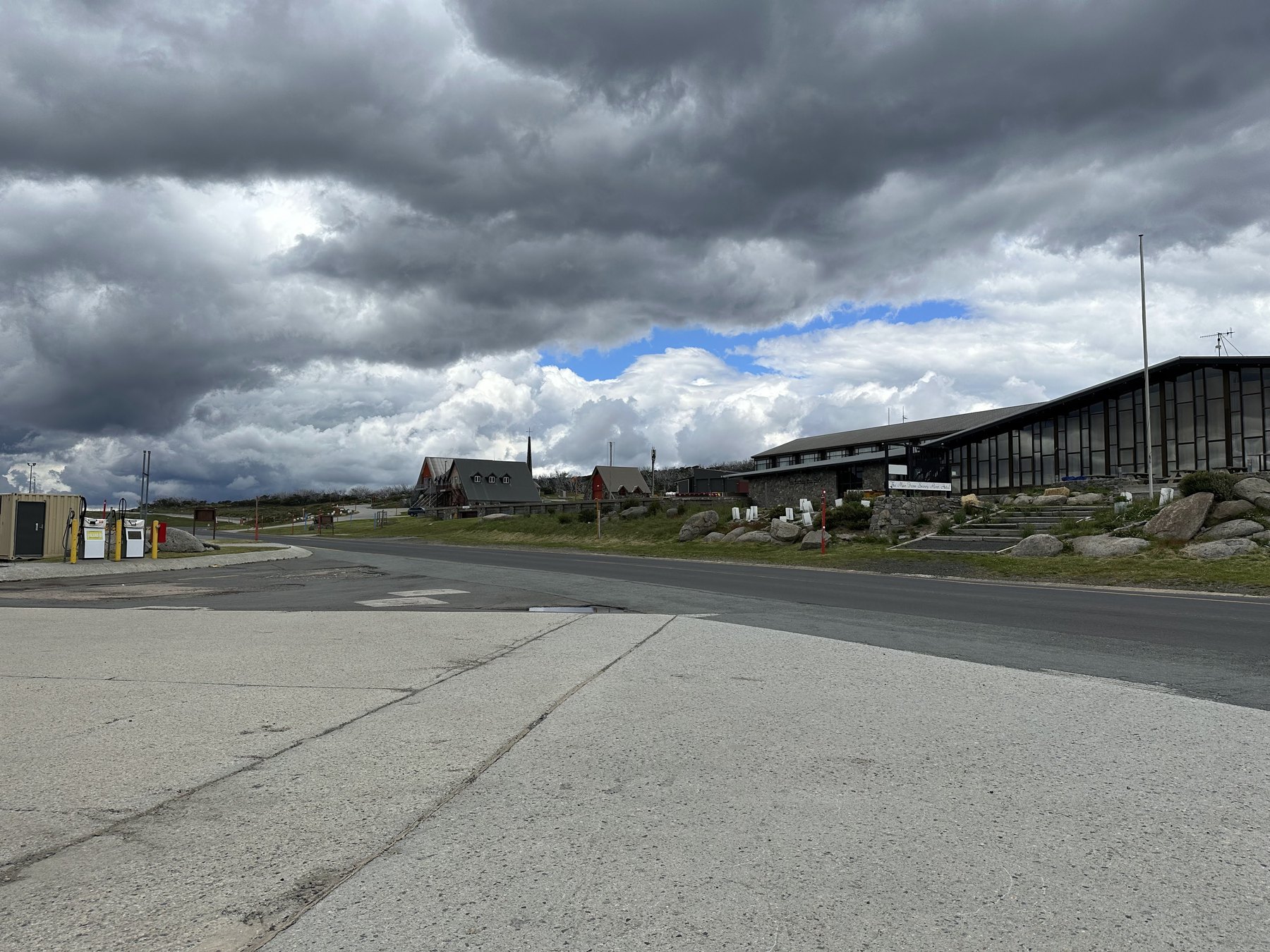 An empty road through Perisher Valley past fuel bowsers, lodges and a long glass-fronted hotel under storm cloud with a patch of blue sky