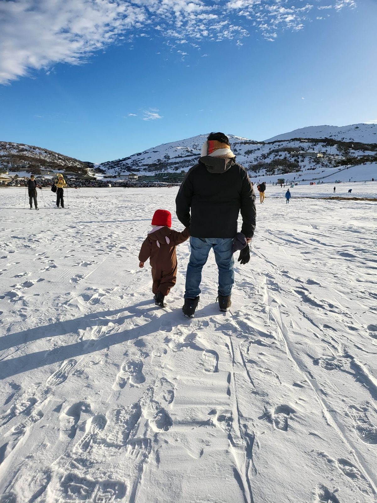 A man and a small child in a snowsuit walking hand in hand away from the camera across deep snow, ski slopes and a blue sky ahead