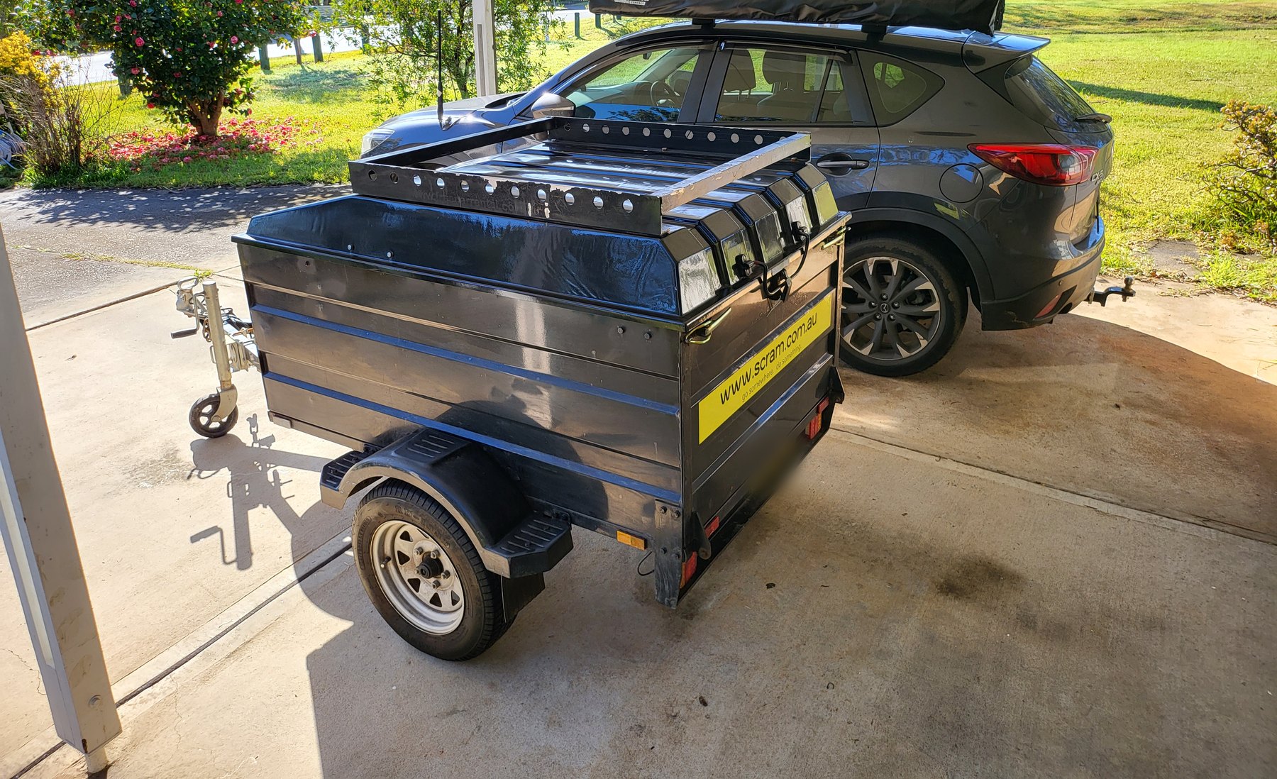 A hired box trailer with a lockable lid and a roof rack on top, parked on a driveway behind a dark grey SUV with a rolled awning on its roof bars