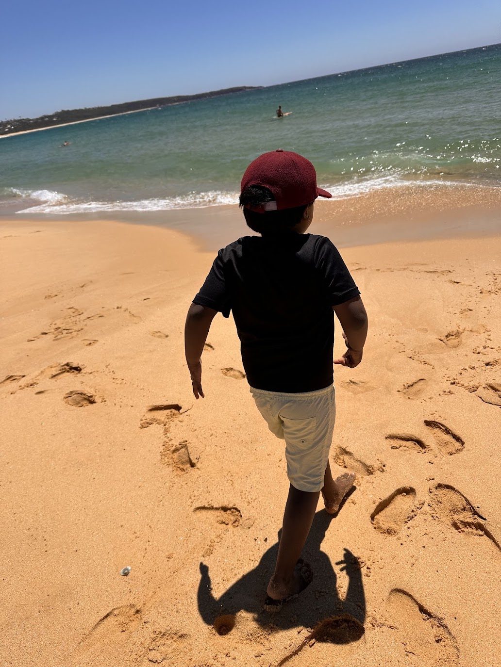 A boy walking down wet sand towards clear green surf, a low headland running out to a dark rocky point on the far side of the beach