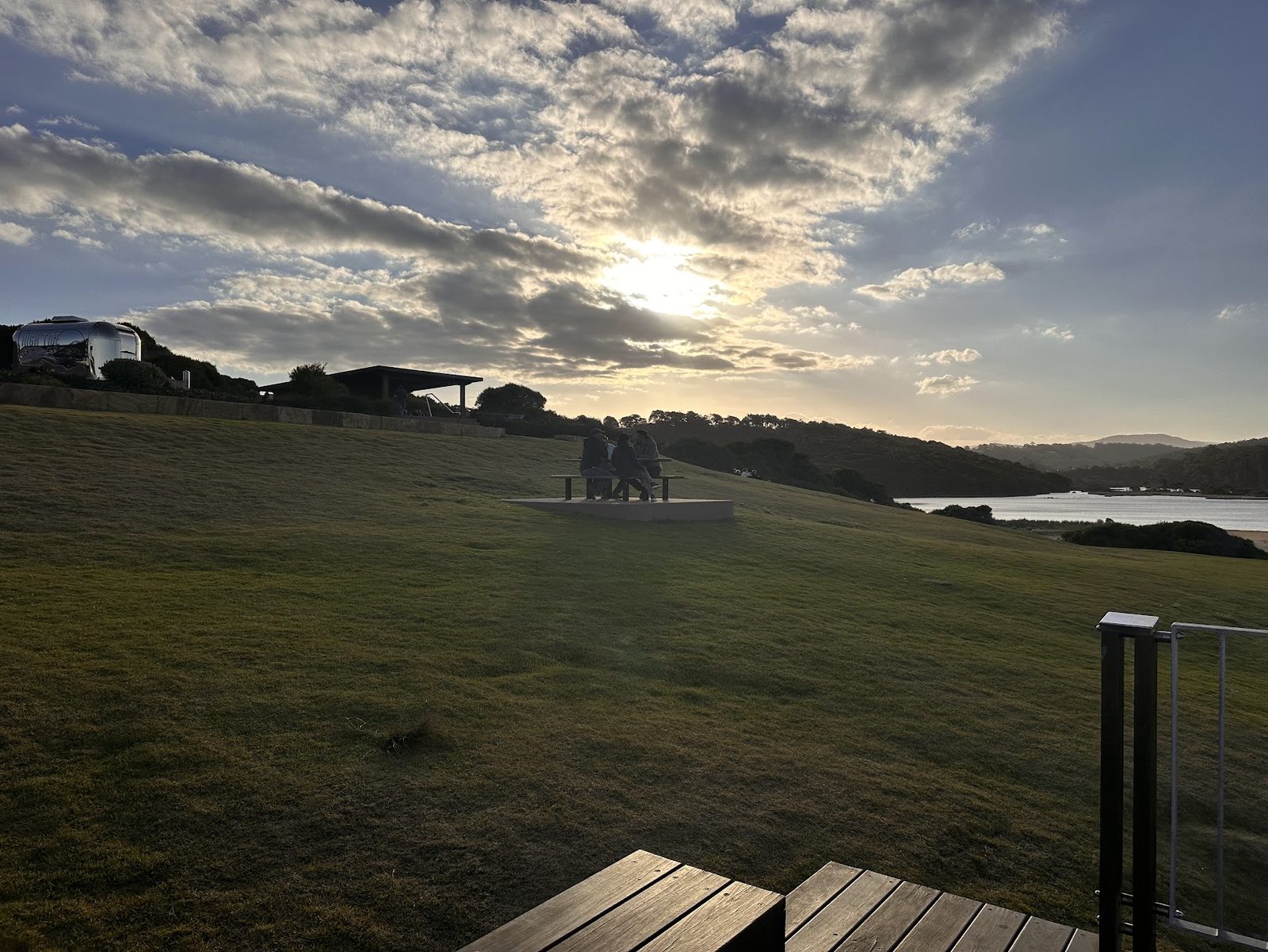 A mown grass slope above an inlet at sunset with a picnic table on a concrete pad, a shelter and a silver Airstream van parked on the rise behind