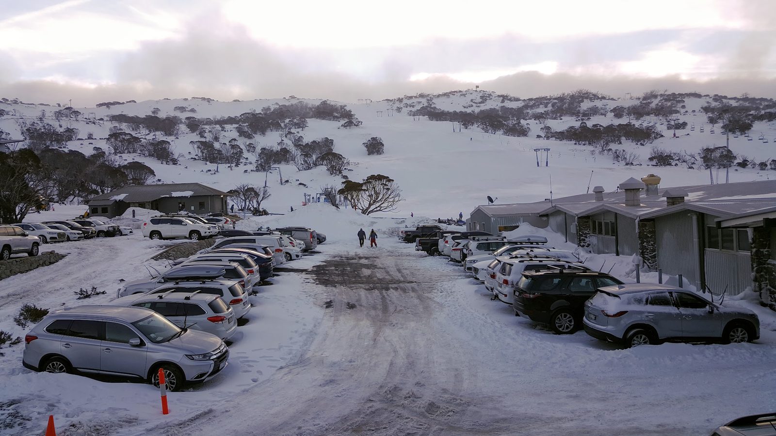 The same car park late in the day under a pale sky, snow-covered cars, two figures walking up the cleared road towards the lifts