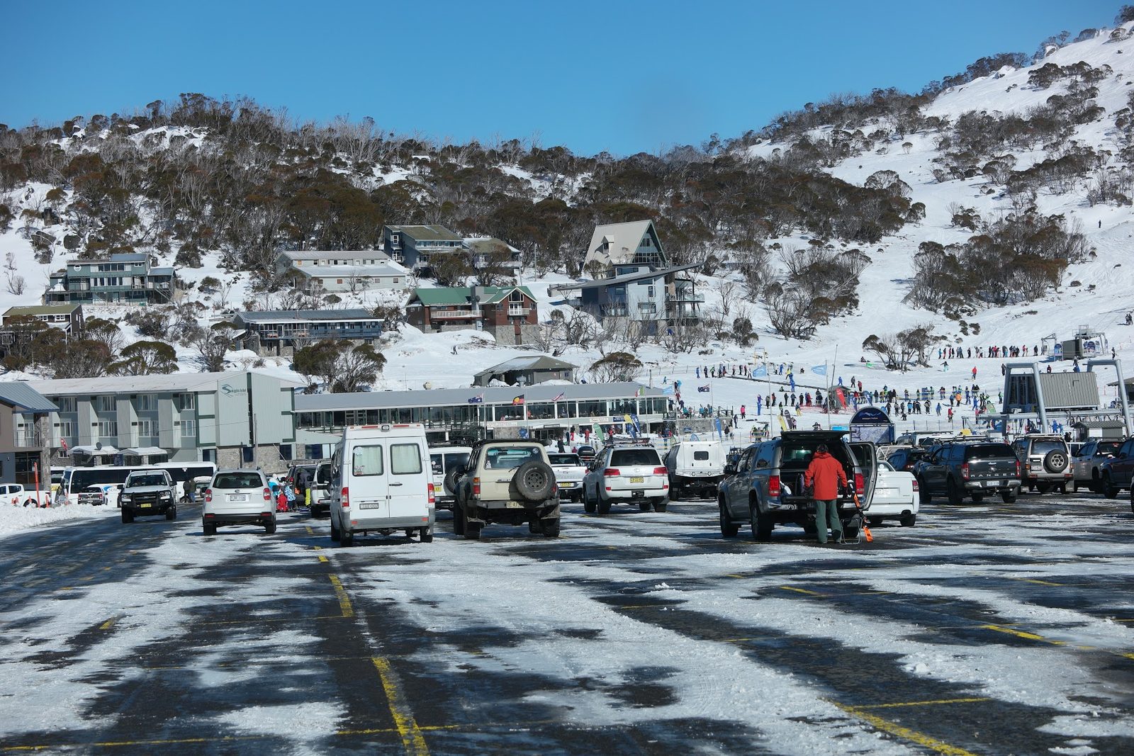 A packed snow car park below a ski village, lodges on the slope above and a beginners area with lift queues off to one side
